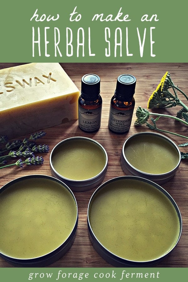 Several tins of homemade herbal salve with a bar of beeswax, essential oils, and herbs in the background.