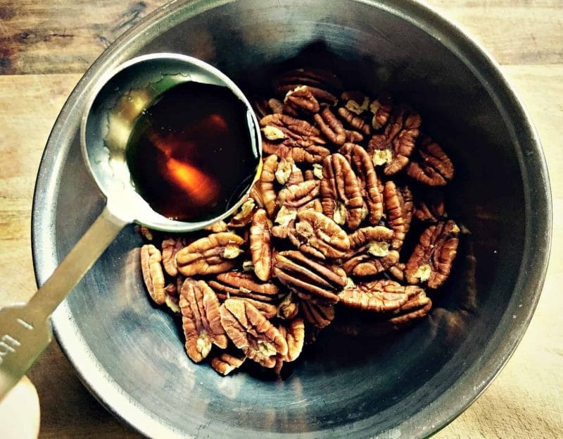 a tablespoon of maple syrup being poured into a bowl of pecans