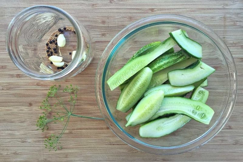 A bowl of cucumbers sliced length wise and a jar of whole spices and garlic cloves at the bottom, with a dill flower laying to the side. On a wood surface.