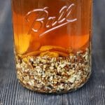 A clear glass jar of dandelion root bitters, the liquid is a burnt orange color and there is chopped up dandelion root at the bottom of the jar.