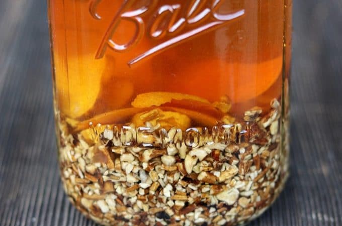 A clear glass jar of dandelion root bitters, the liquid is a burnt orange color and there is chopped up dandelion root at the bottom of the jar.