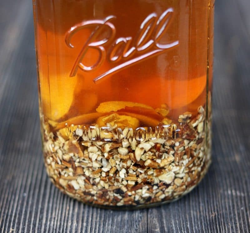 A clear glass jar of dandelion root bitters, the liquid is a burnt orange color and there is chopped up dandelion root at the bottom of the jar. 