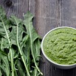 A bowl of dandelion pesto next to a pile of dandelion greens on a wood surface.