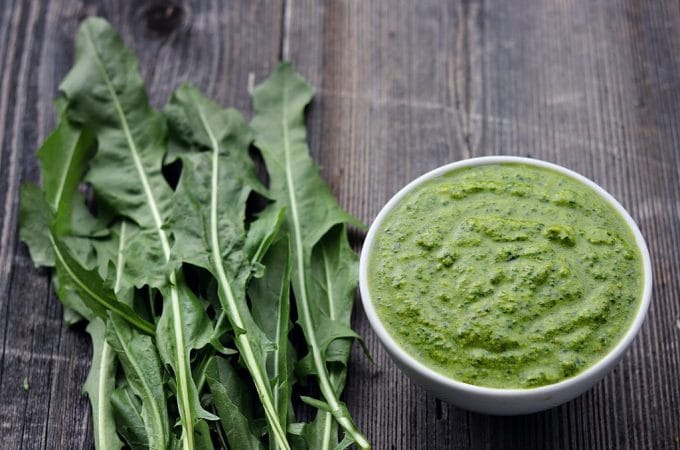 A bowl of dandelion pesto next to a pile of dandelion greens on a wood surface.