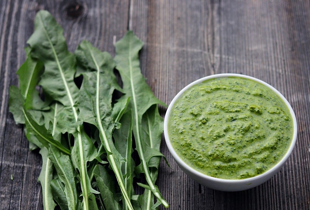 A bowl of dandelion pesto next to a pile of dandelion greens on a wood surface.