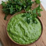 A bowl of chickweed pesto with fresh chickweed surrounding on a wood cutting board, top view.