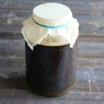 Root beer fermenting in a large jar with cheesecloth on top, on a gray wood surface.