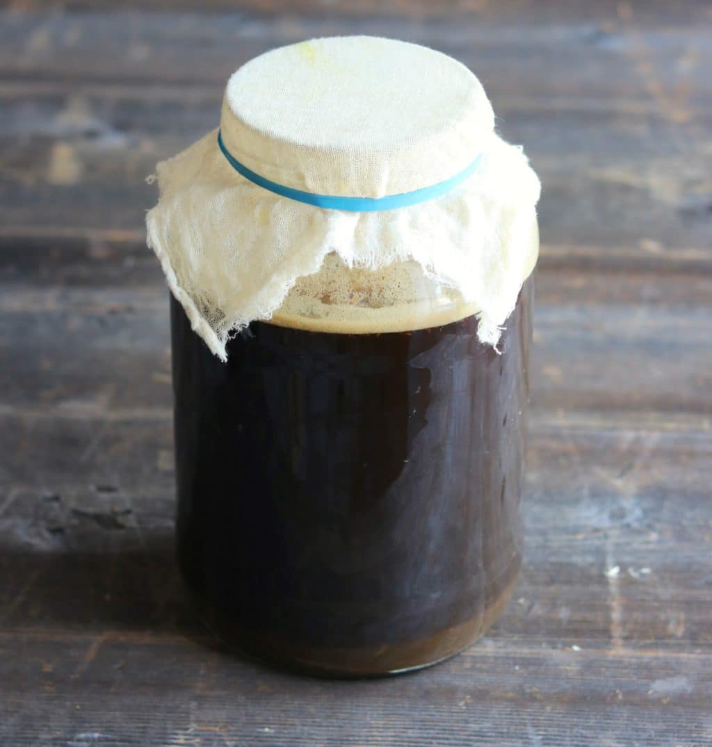 Root beer fermenting in a large jar with cheesecloth on top, on a gray wood surface. 