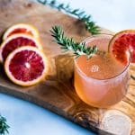 A rosemary blood orange champagne cocktail in a stemless wineglass, with a sprig of rosemary in it and a blood orange slice on the rim.