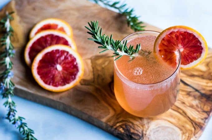 A rosemary blood orange champagne cocktail in a stemless wineglass, with a sprig of rosemary in it and a blood orange slice on the rim.