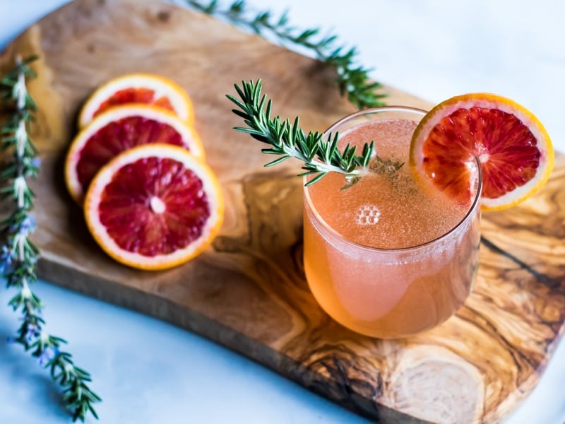 A rosemary blood orange champagne cocktail in a stemless wineglass, with a sprig of rosemary in it and a blood orange slice on the rim. 