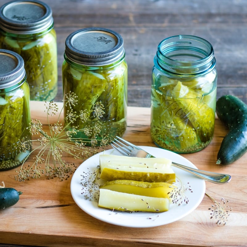 jars of homemade kosher dill pickles on a wooden cutting board with a small white plate of pickles