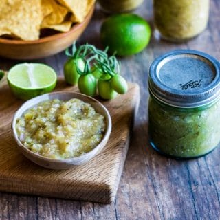 A jar of green tomato salsa verde next to a wood cutting board with a small white bowl filled with the green salsa, surrounded by limes, green tomatoes, and tortilla chips.