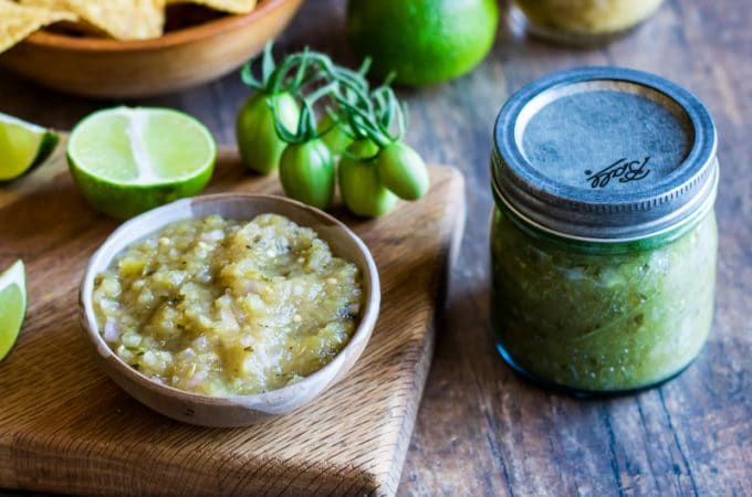 A jar of green tomato salsa verde next to a wood cutting board with a small white bowl filled with the green salsa, surrounded by limes, green tomatoes, and tortilla chips.