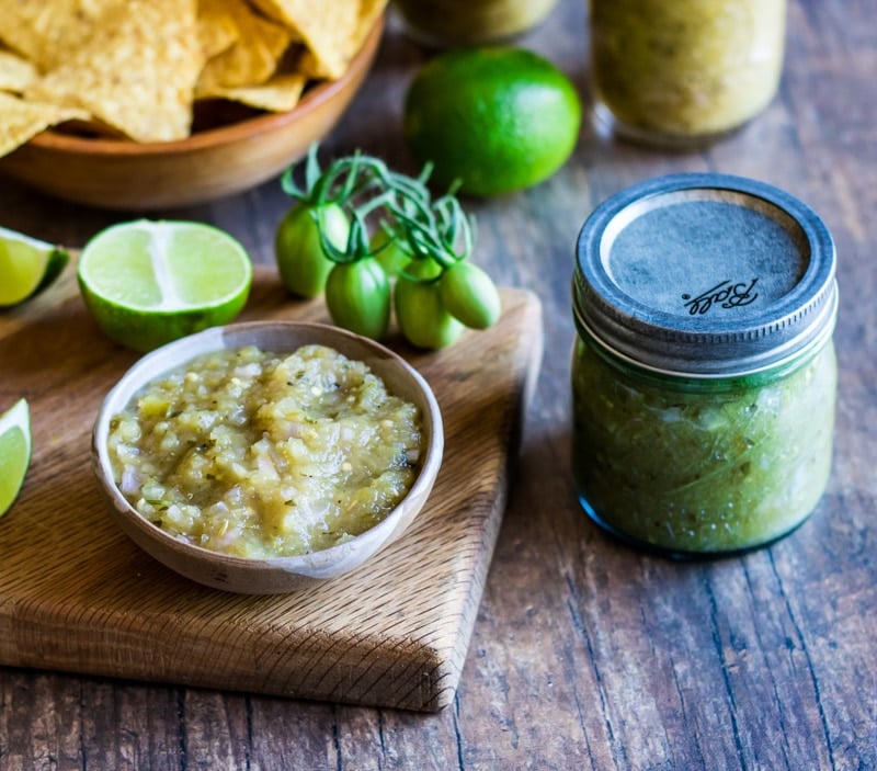 A jar of green tomato salsa verde next to a wood cutting board with a small white bowl filled with the green salsa, surrounded by limes, green tomatoes, and tortilla chips.  