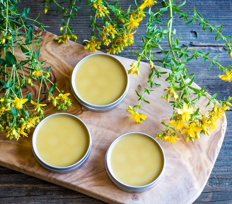 St. John's wort salve in tins, on a wooden cutting board, surrounded by fresh St. John's wort flowers and stems. 