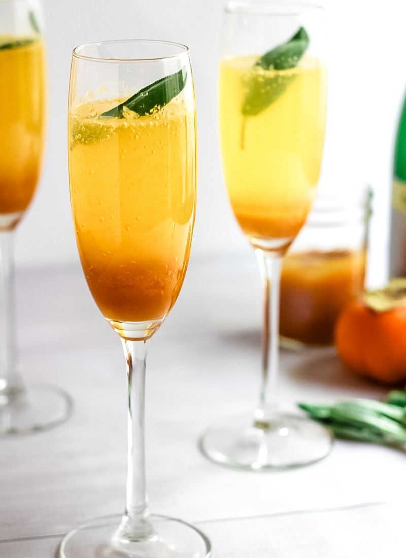 Several champagne flutes with persimmon champagne cocktail, which is a darker orange at the bottom, a lighter orange on top, and each garnished with a fresh sage leaf.