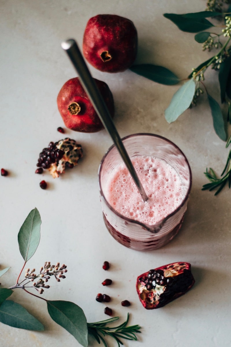 A clear glass pitcher of pomegranate martini being stirred, top view, surrounded by a pomegranate, pomegranate seeds, and fresh leaves. 