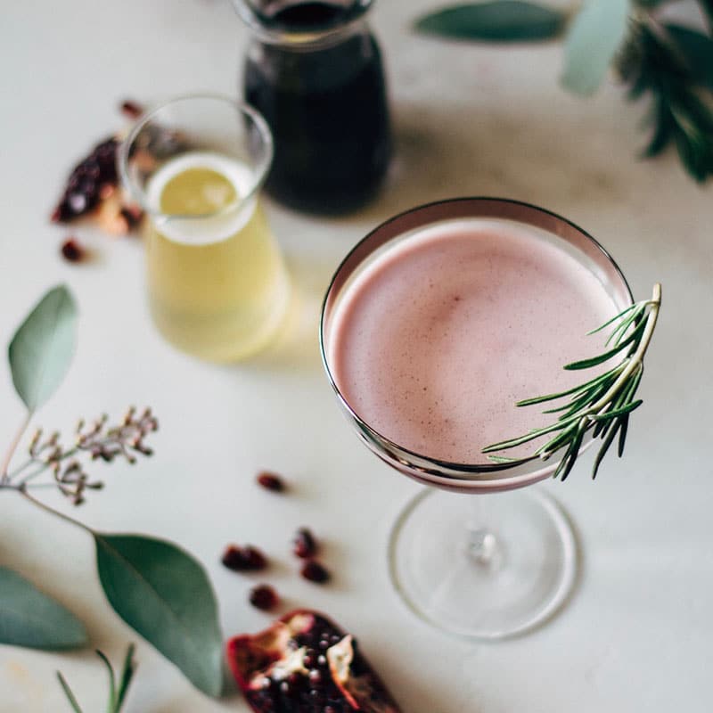 A pomegranate martini top view in a glass with a rosemary sprig, surrounded by a glass of rosemary syrup, pomegranate seeds, and fresh leaves. 