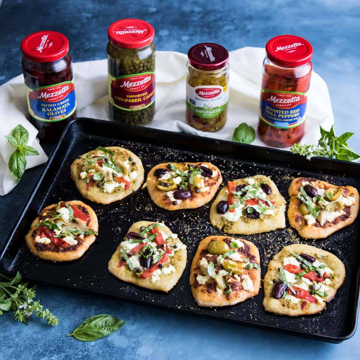 mini sourdough pizzas on a sheet pan with jars of Mezzetta ingredients