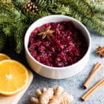 A white bowl of cranberry sauce on a countertop surrounded by cinnamon sticks, orange slices, ginger, and evergreen branches.