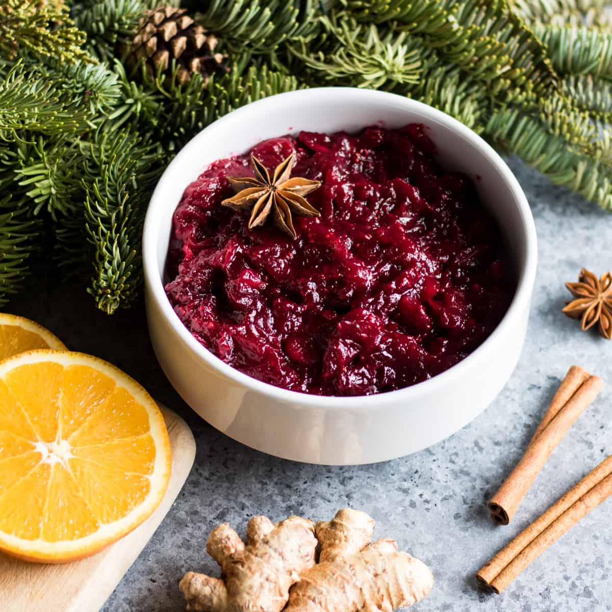 A white bowl of cranberry sauce on a countertop surrounded by cinnamon sticks, orange slices, ginger, and evergreen branches. 