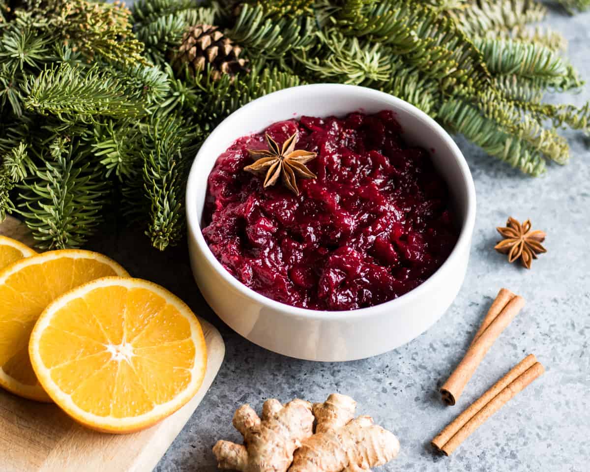 a bowl of spiced cranberry sauce on a table with whole spices, orange slices, and a conifer branch