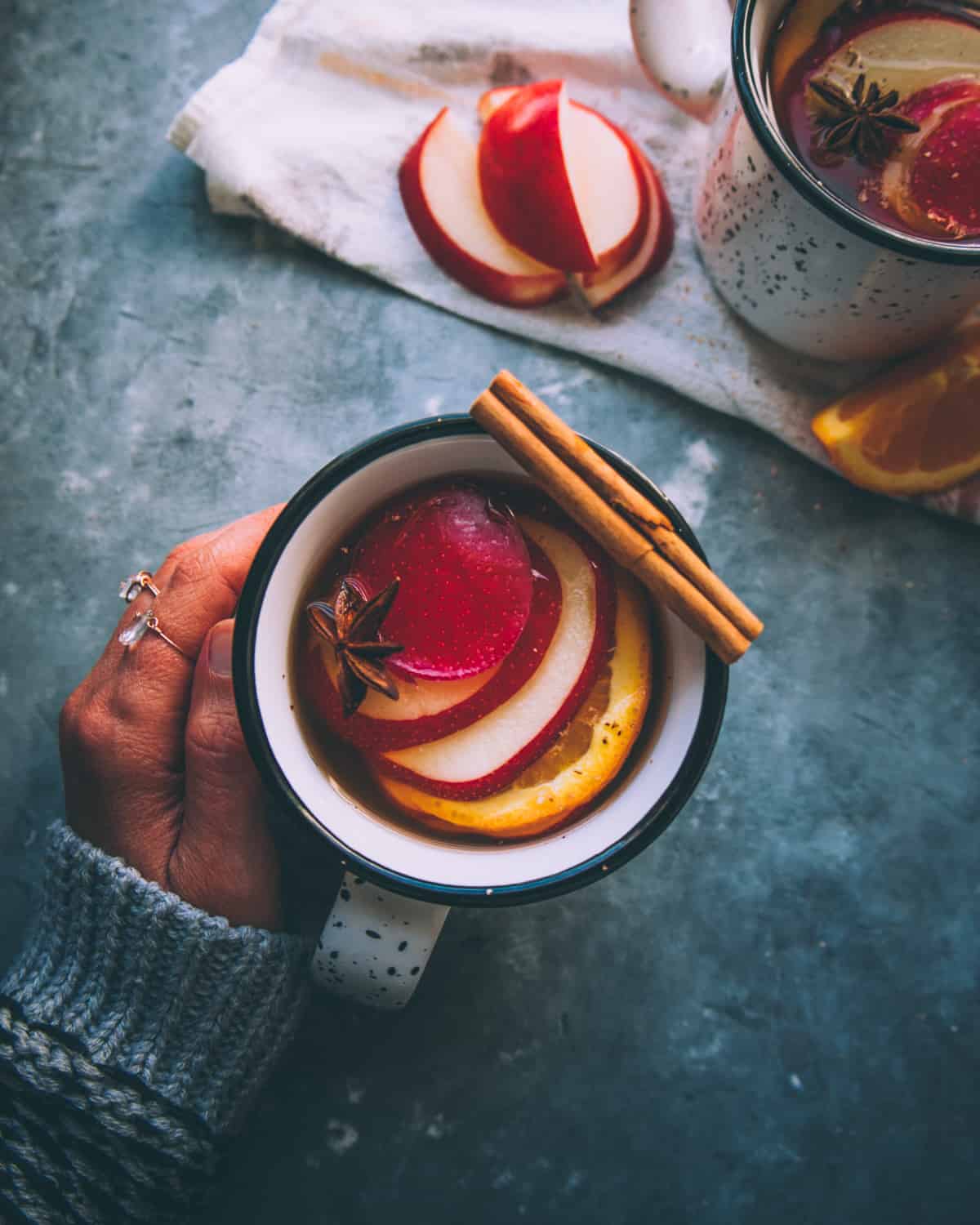 a hand holding a mug of mulled hard cider garnished with apple slices and a cinnamon stick