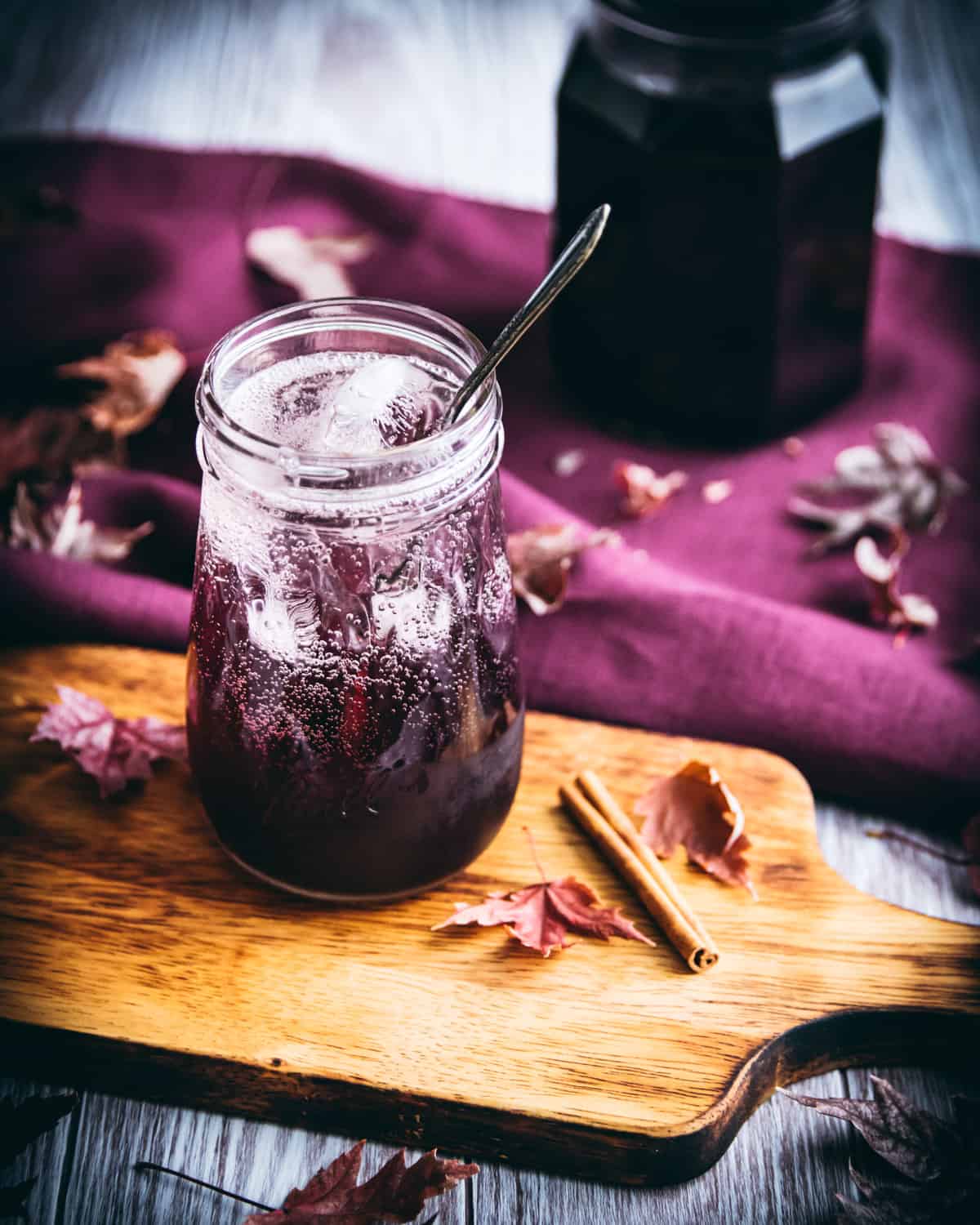 An elderberry cocktail in a clear jar glass with ice and a spoon, sitting on a wooden cutting board. Surrounded by fall leaves, cinnamon, and a jar of elderberry syrup. 