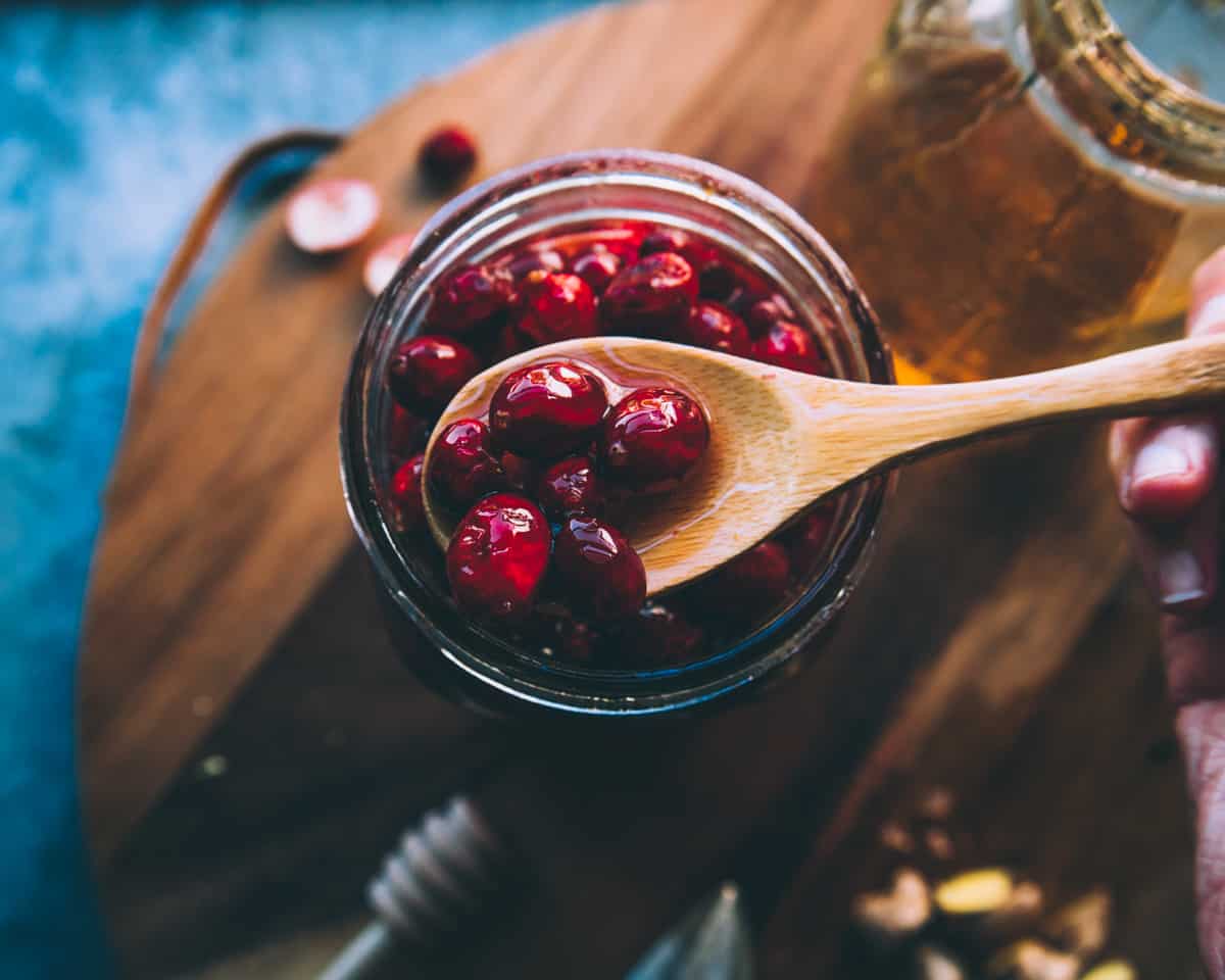 A jar of honey fermented cranberries with a wooden spoon lifting some out, top view. On a dark wood surface. 