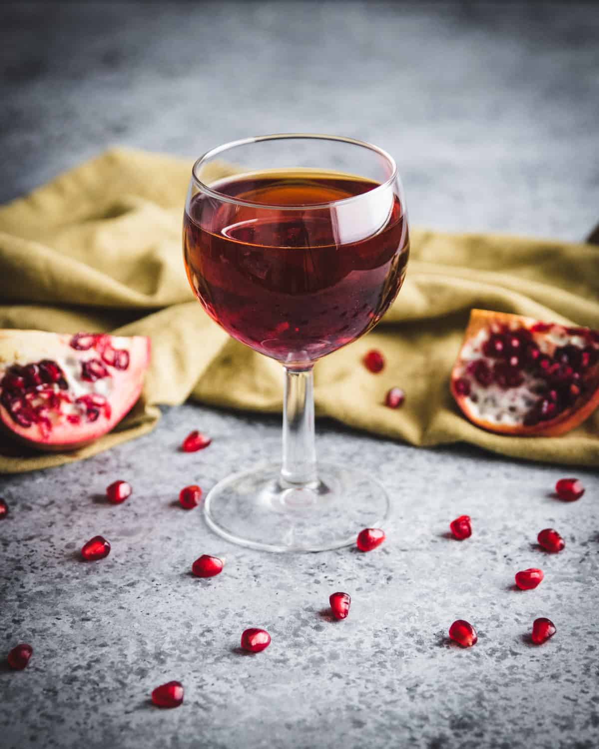 A wine glass with red pomegranate wine, surrounded by an open pomegranate and pomegranate seeds.