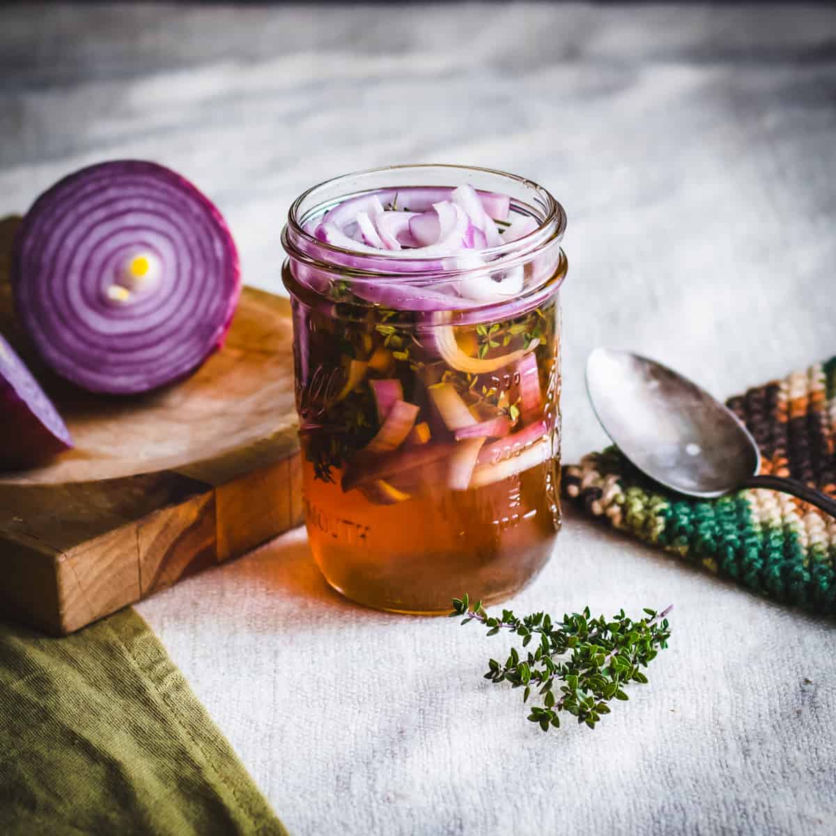 A jar of fermented red onion and thyme in honey on a white cloth surrounded by half of a red onion and fresh thyme.