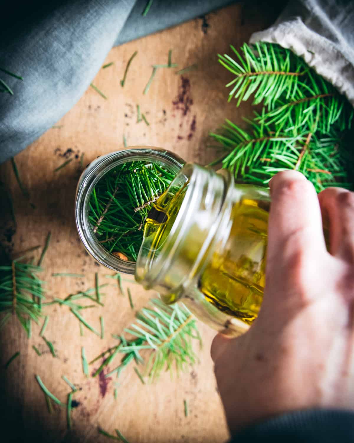 a hand pouring olive oil into a jar of conifer needles