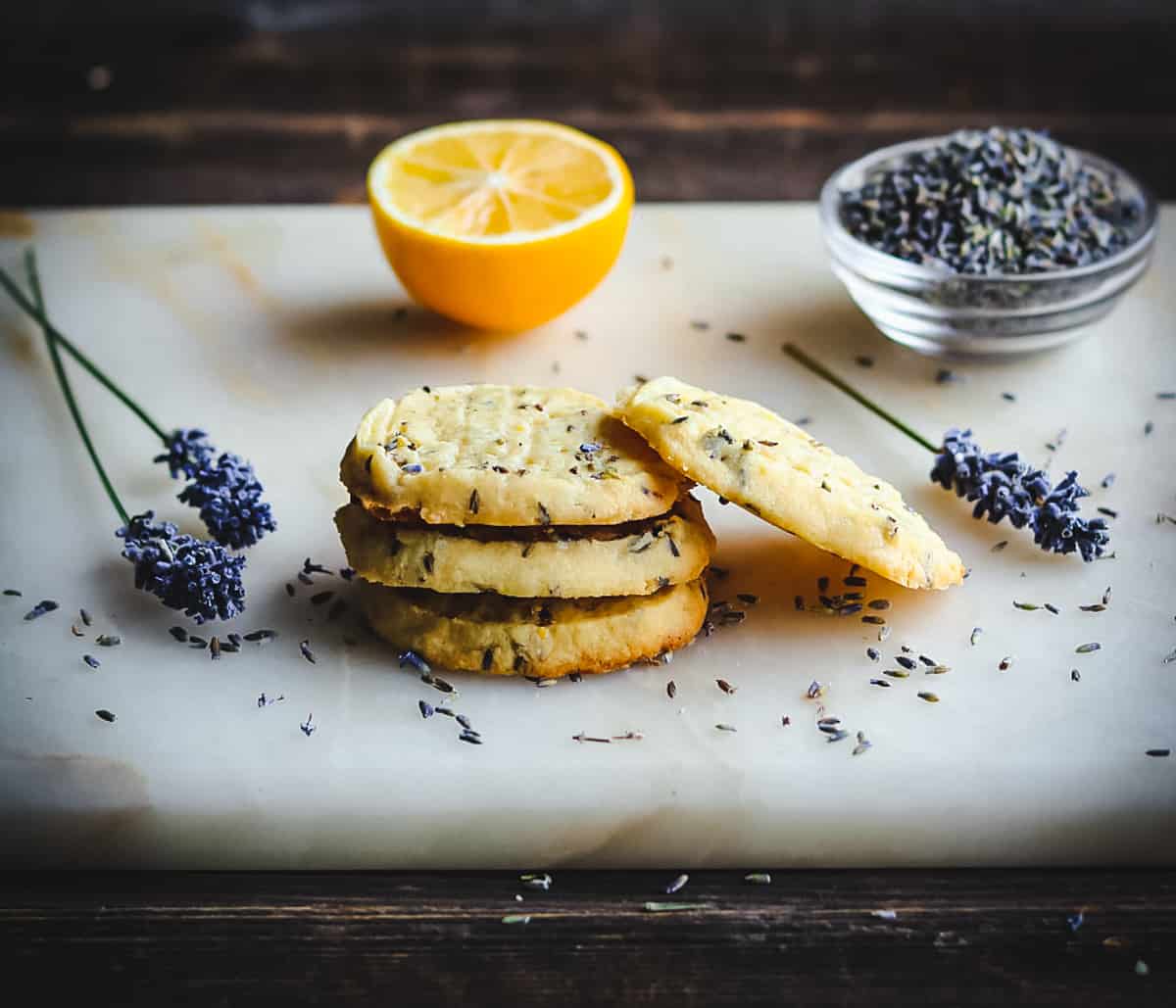 A stack of lemon lavender shortbread cookies on a board with lavender flowers and a halved lemon.