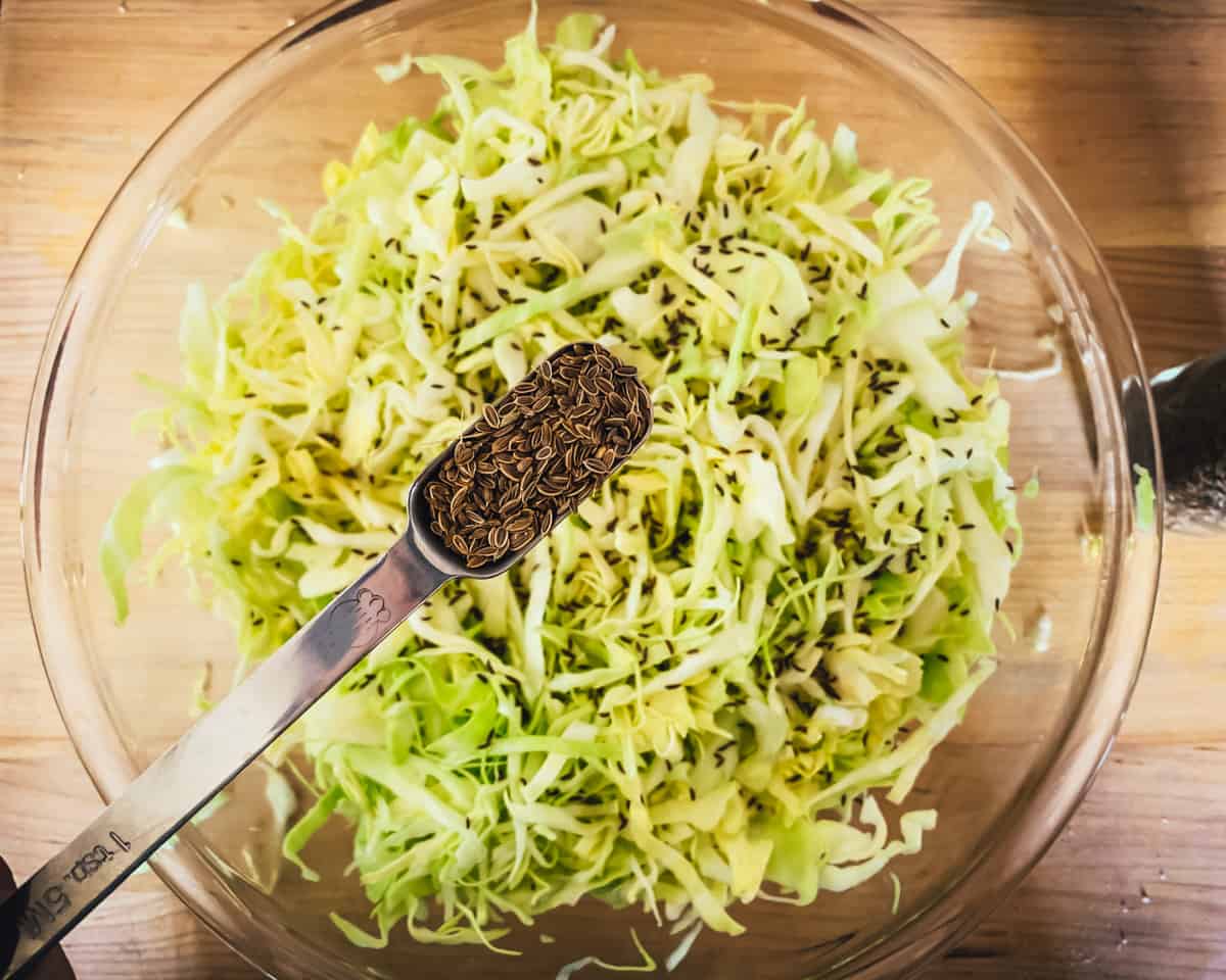 a tablespoon of dill seeds being added to the cabbage