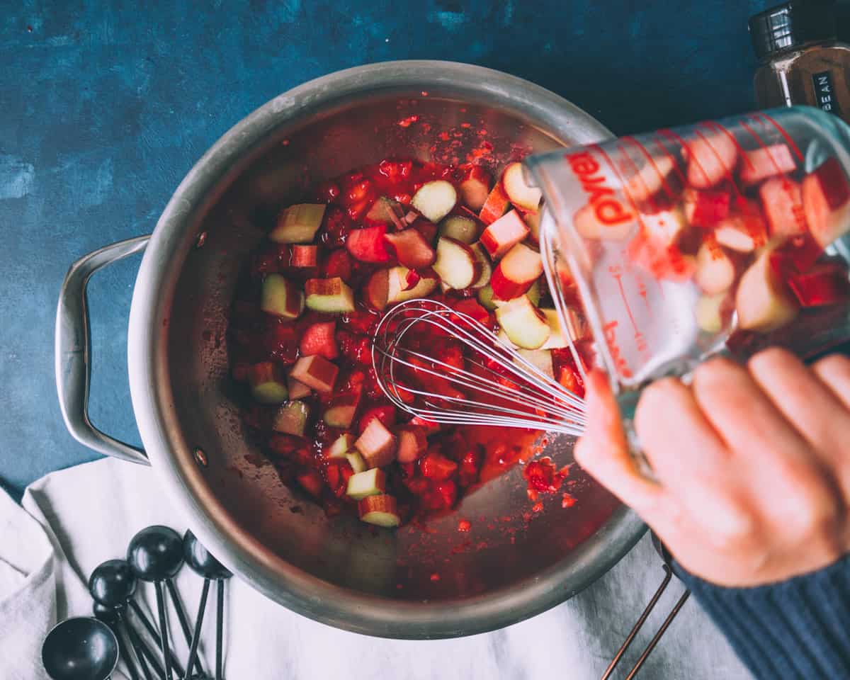 a woman's hand adding rhubarb to a pot with strawberries