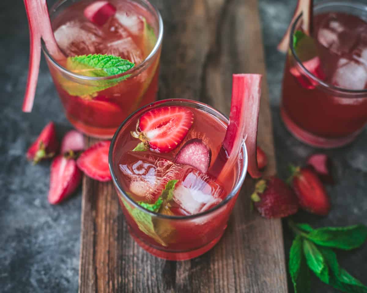 three glasses of fermented strawberry rhubarb soda on a wooden board garnished with a rhubab stalk
