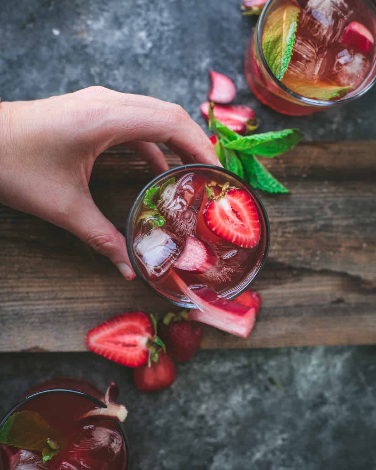 Glasses of strawberry rhubarb soda on a wood surface with a hand holding one, garnished with fresh strawberries and a rhubarb stalk. 