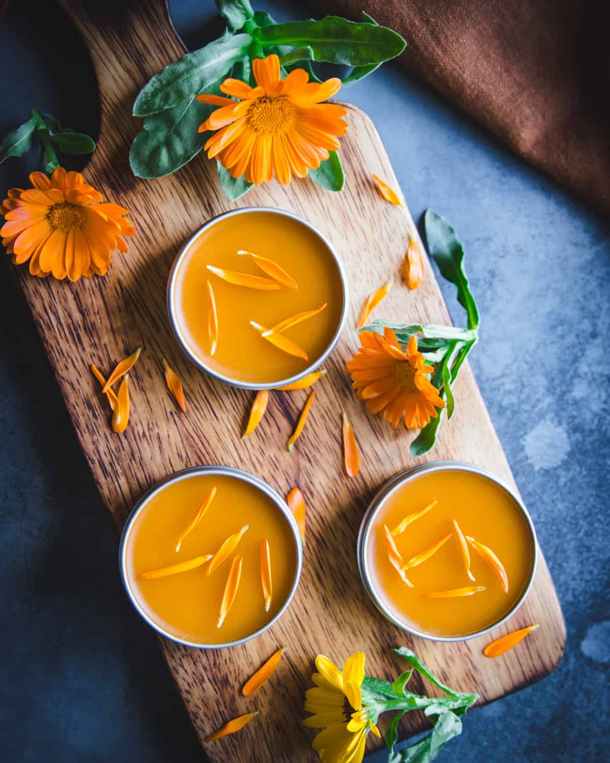 3 tins of calendula salve on a wooden cutting board, surrounded by calendula flowers. 