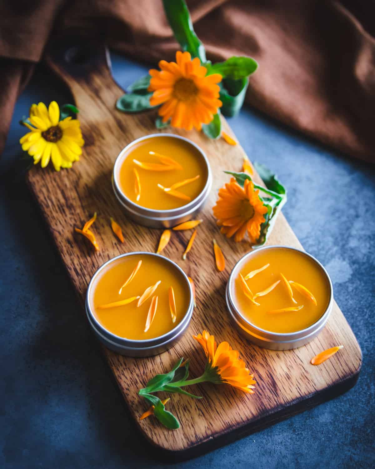 three tins of calendula salve on a wooden cutting board with calendula flowers