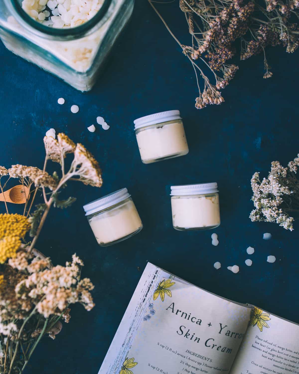 3 small jars with lids on, resting on their sides and filled with arnica and yarrow cream. On a navy blue countertop surrounded by the recipe book open to the page showing arnica and yarrow skin cream recipe, and dried yarrow flowers.