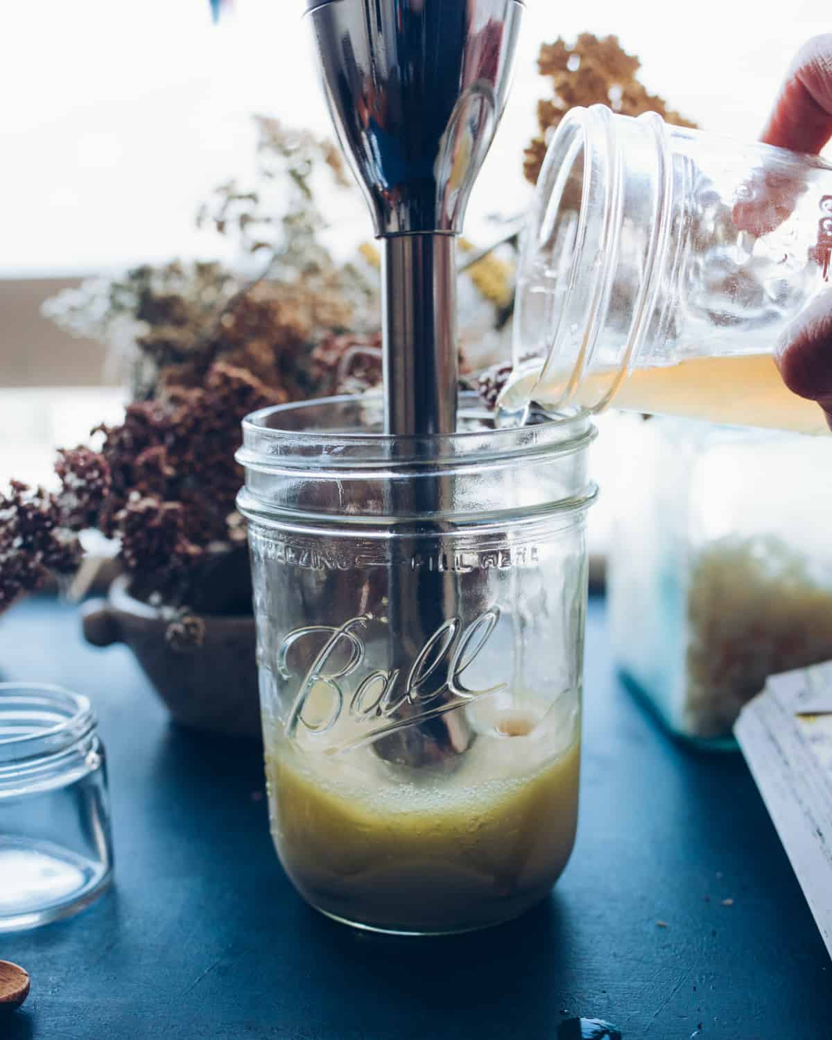 A clear wide mouth jar with liquid from the recipe being blended with an immersion blender as it pours in. On a dark blue counter with dried yarrow flowers in background. 