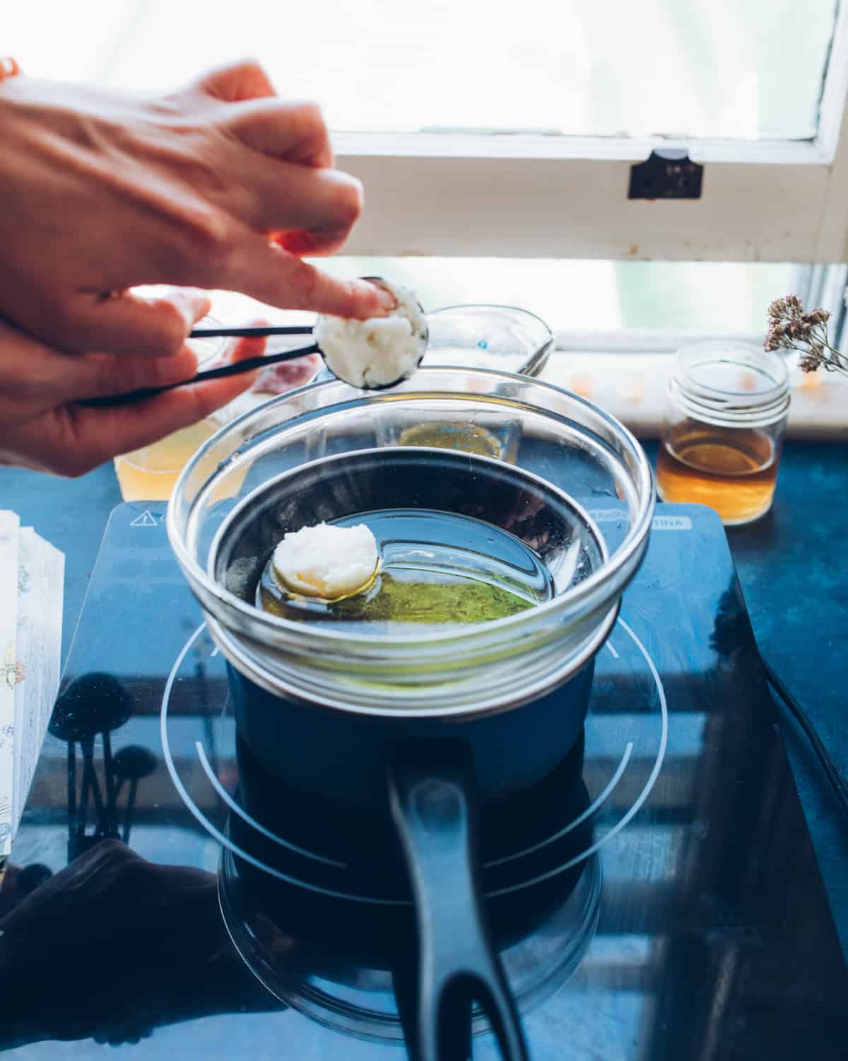 A pot on a black glass stove top, with a clear glass bowl atop being used as a double boiler. Bowl is half filled with infused oil, with mango butter being dropped into bowl with a tablespoon by hand. 