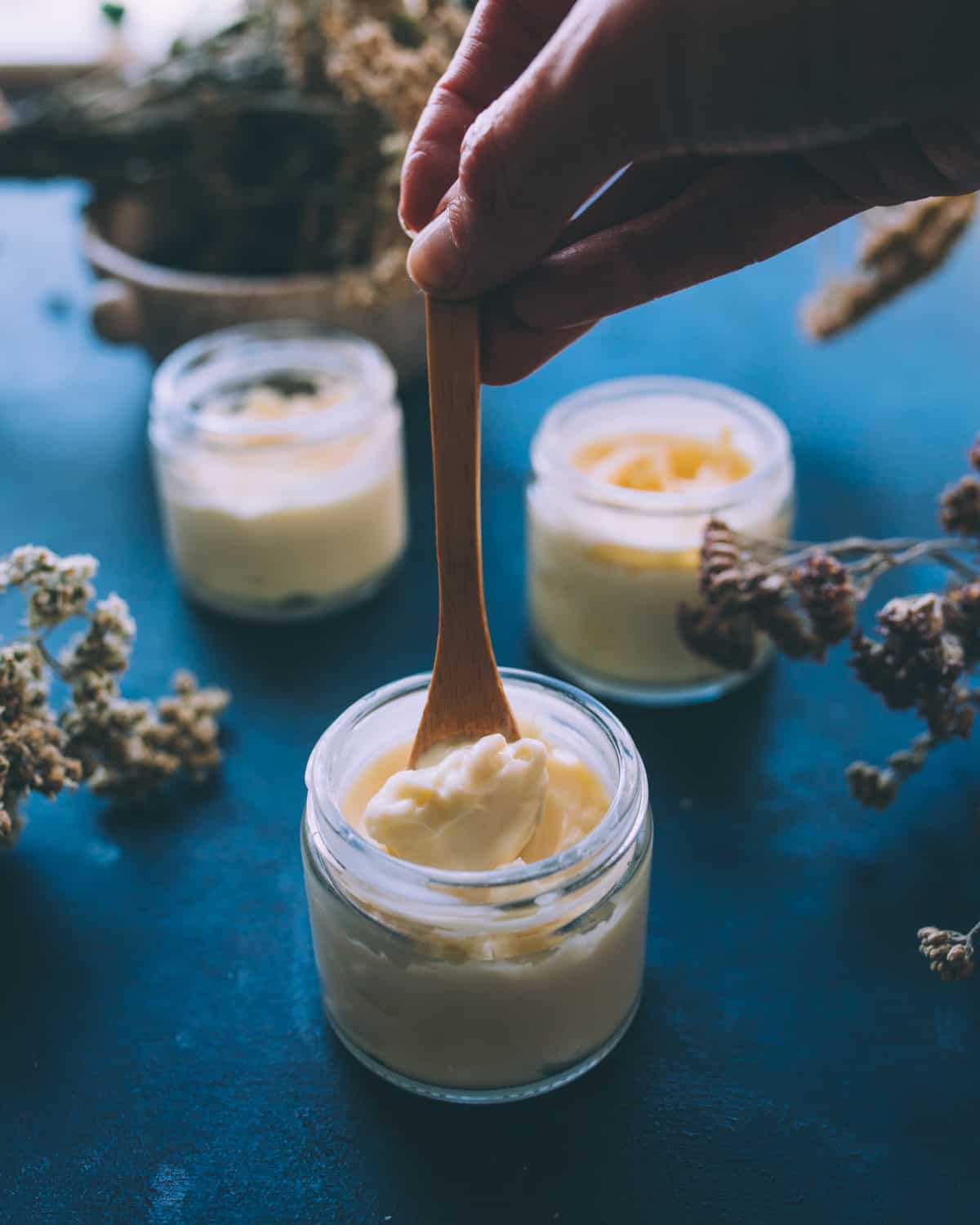 3 jars with no lids, filled with arnica and yarrow cream. The jar in the front has a small wooden spoon scooping out some cream to show the thick creamy texture, the other two jars are in the background on a dark blue countertop with dried yarrow flowers. 