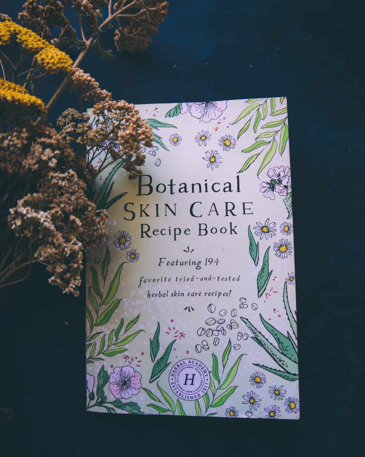 Dark background featuring a light purple book cover with darker purple flowers and their green leaves surrounding the book title that reads "Botanical Skin Care Recipe Book featuring 194 favorite tried-and-tested herbal skincare recipes" 