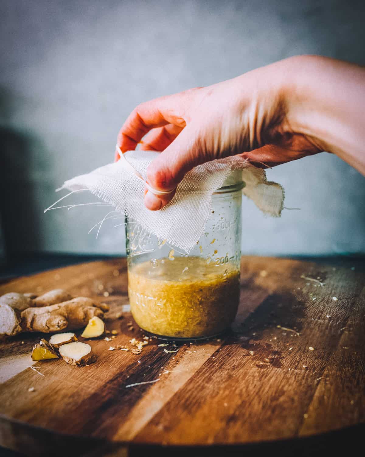 a womans hand covering the jar of ginger bug with a cheesecloth and rubber band