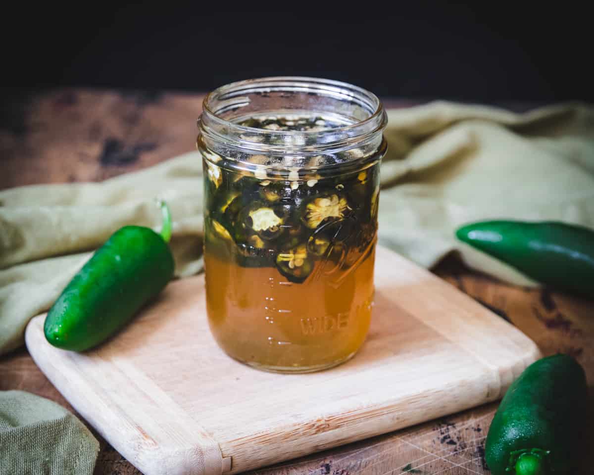 Atop a wooden cutting board sits a jar of fermented jalapeños in honey, and fresh whole jalapeños in the background. 