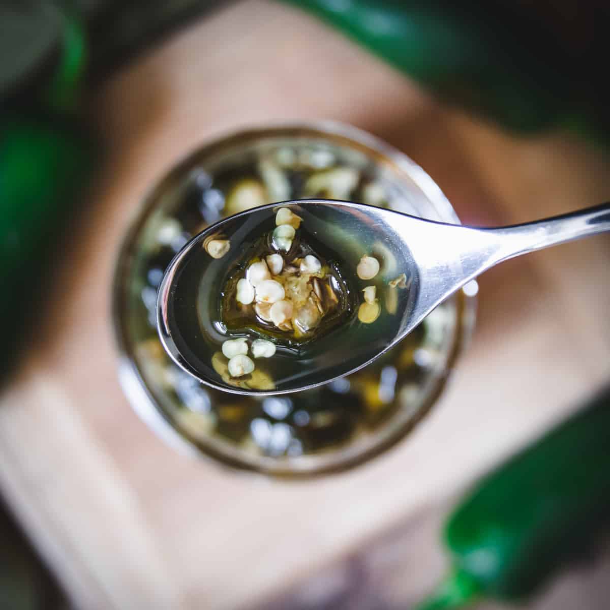 Spoon held above a jar, both filled with honey and bits of chopped jalapeños, with a wooden cutting board in background.