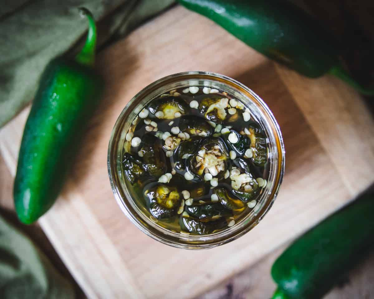 Chopped jalapenos in a jar filled with honey, on a wood cutting board with a fresh jalapeño in the background.