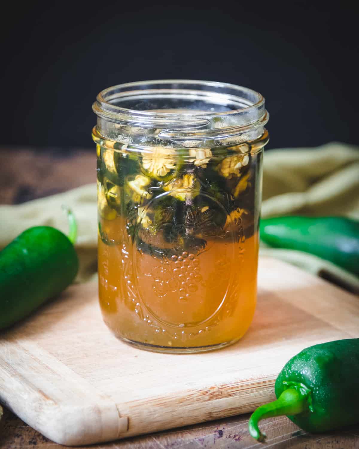 Fermented sliced jalapeños floating at the top of a jar of honey, on a wood cutting board with whole fresh jalapeños in the background. 